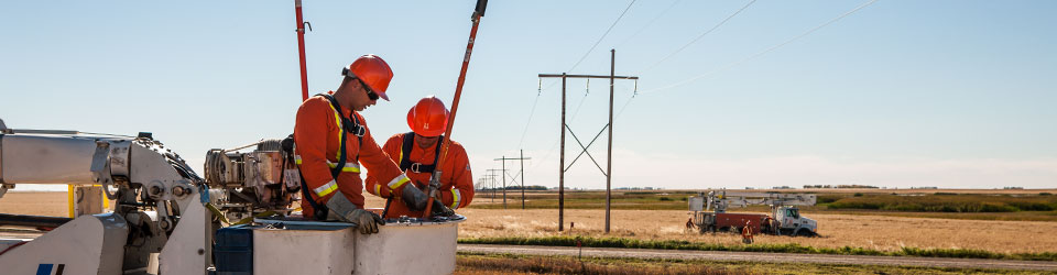 Two power line technicians in a bucket truck with a SaskPower truck and power line in the distance. 